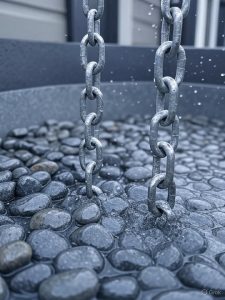 A close-up of a galvanized steel coil rain chain with water running down its links from a gutter into a basin of river stones.